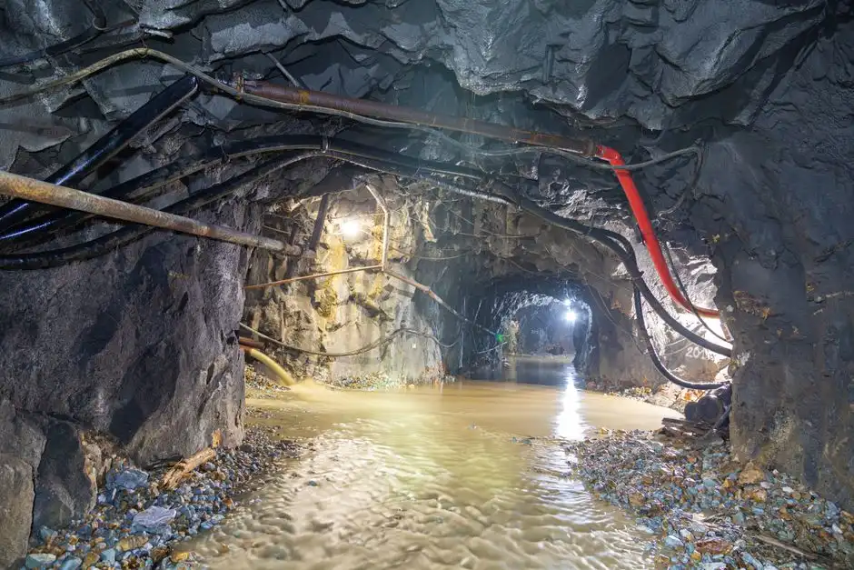 Dimly lit underground cave with flowing water and exposed pipes, showcasing rugged geology.